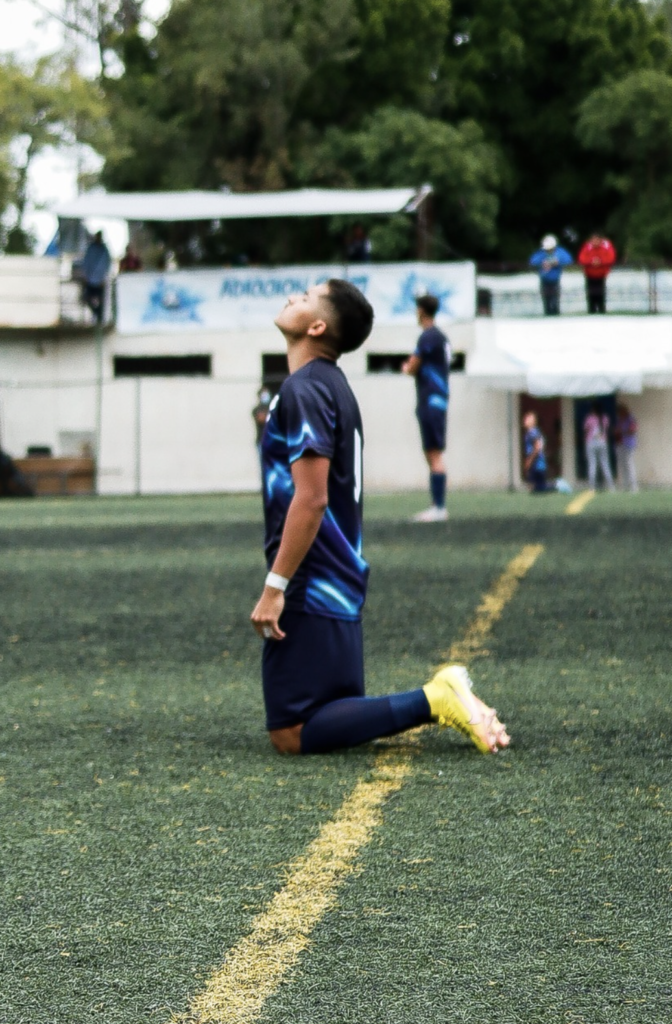 Fotografía deportiva de joven de rodillas con uniforme azul en campo de fútbol de pasto sintético