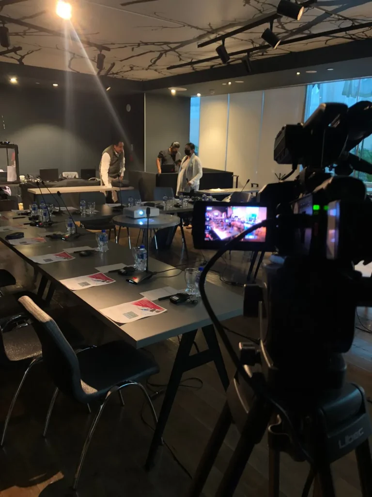 Cámara profesional en trípode enfocando una sala de conferencias en preparación para un streaming en vivo. En la mesa hay micrófonos, documentos y botellas de agua, mientras el equipo técnico ajusta detalles en el fondo