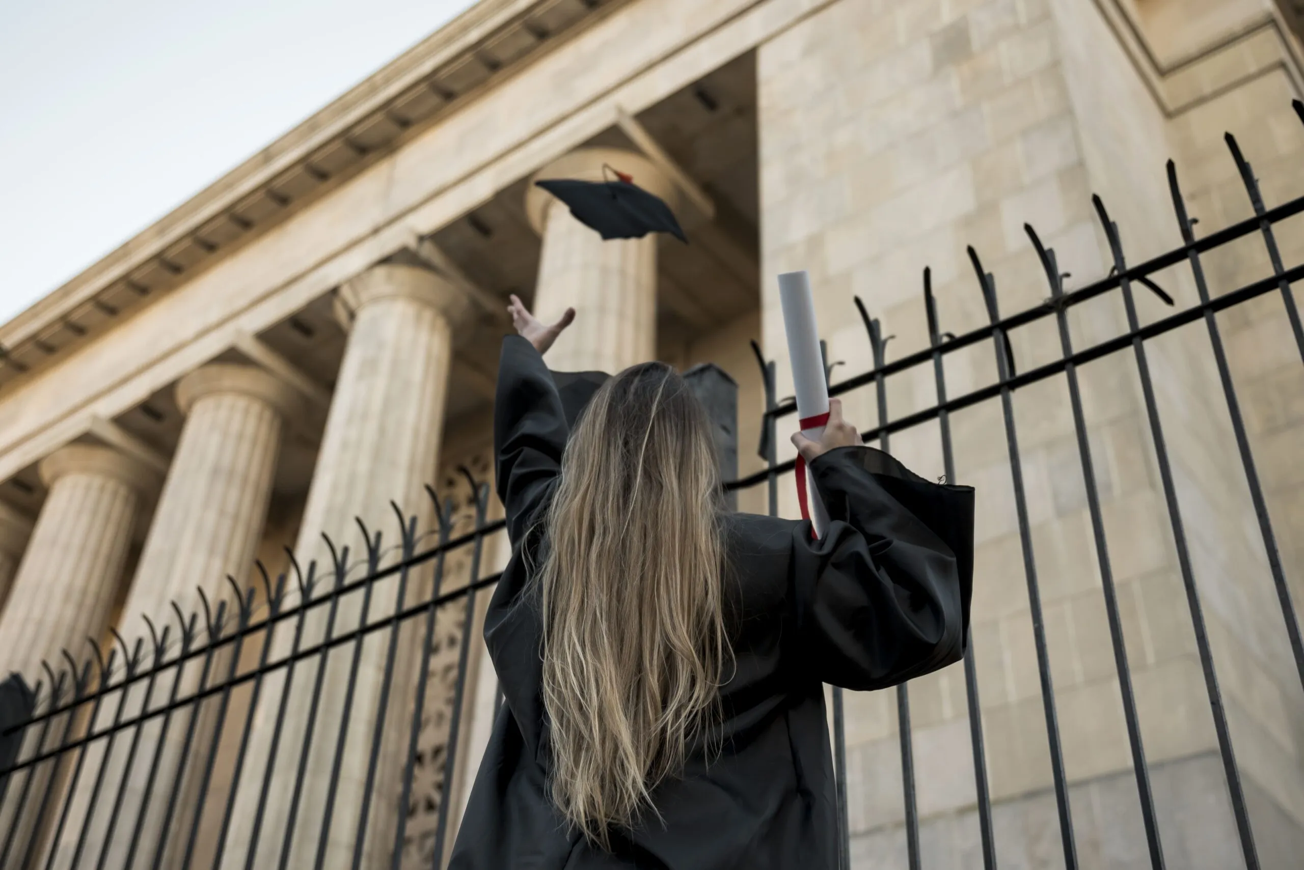Chica lanzando su birrete al aire para su foto de graduación frente a edificio Fotografía de una chica rubia lanzando su birrete al aire para su foto de graduación frente a edificio